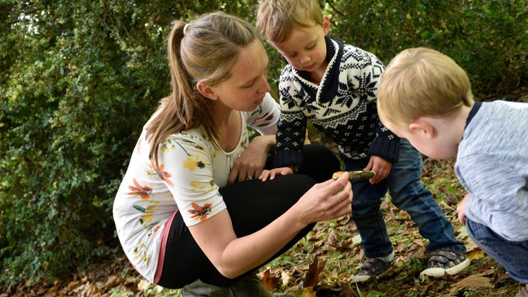 A parent and two young children play with sticks and autumn leaves.